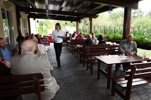 A covered outdoor dining area with wooden benches and tables, a waiter walking through, and people seated and eating. - Dolce Villa Hotel (Prague)