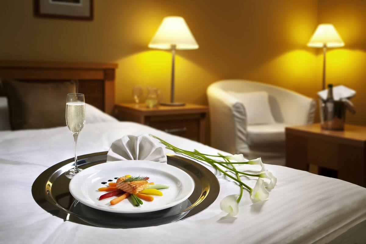 A neatly made bed with a tray of food, a glass of champagne, and a bouquet of flowers.