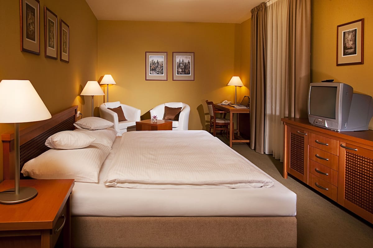 A hotel room with a large bed, two bedside tables with lamps, a desk with a chair, a television, and framed pictures on the wall.