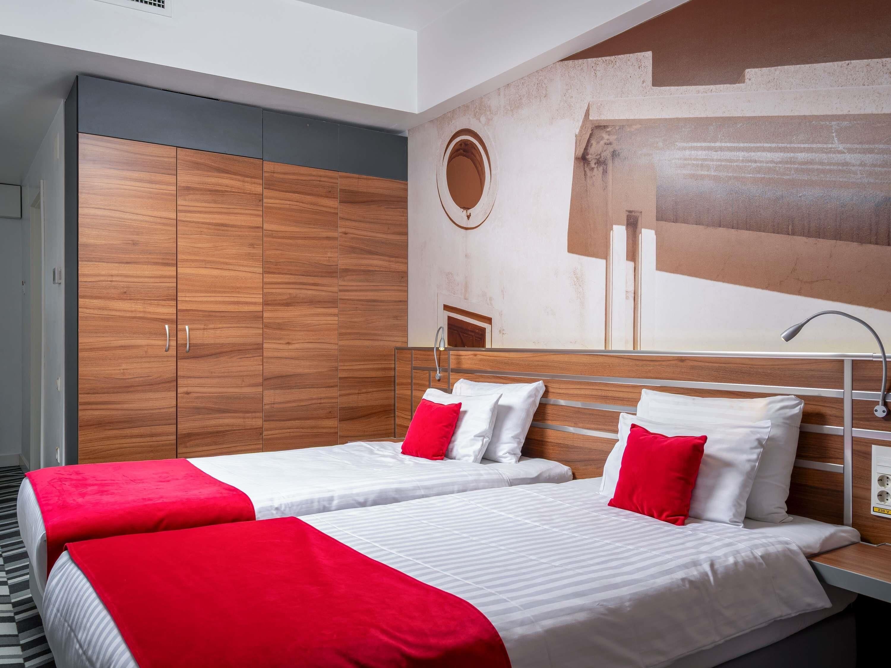 Hotelkamer met twee bedden, elk voorzien van wit beddengoed en rode kussens, een houten garderobe en een modern wandontwerp.
