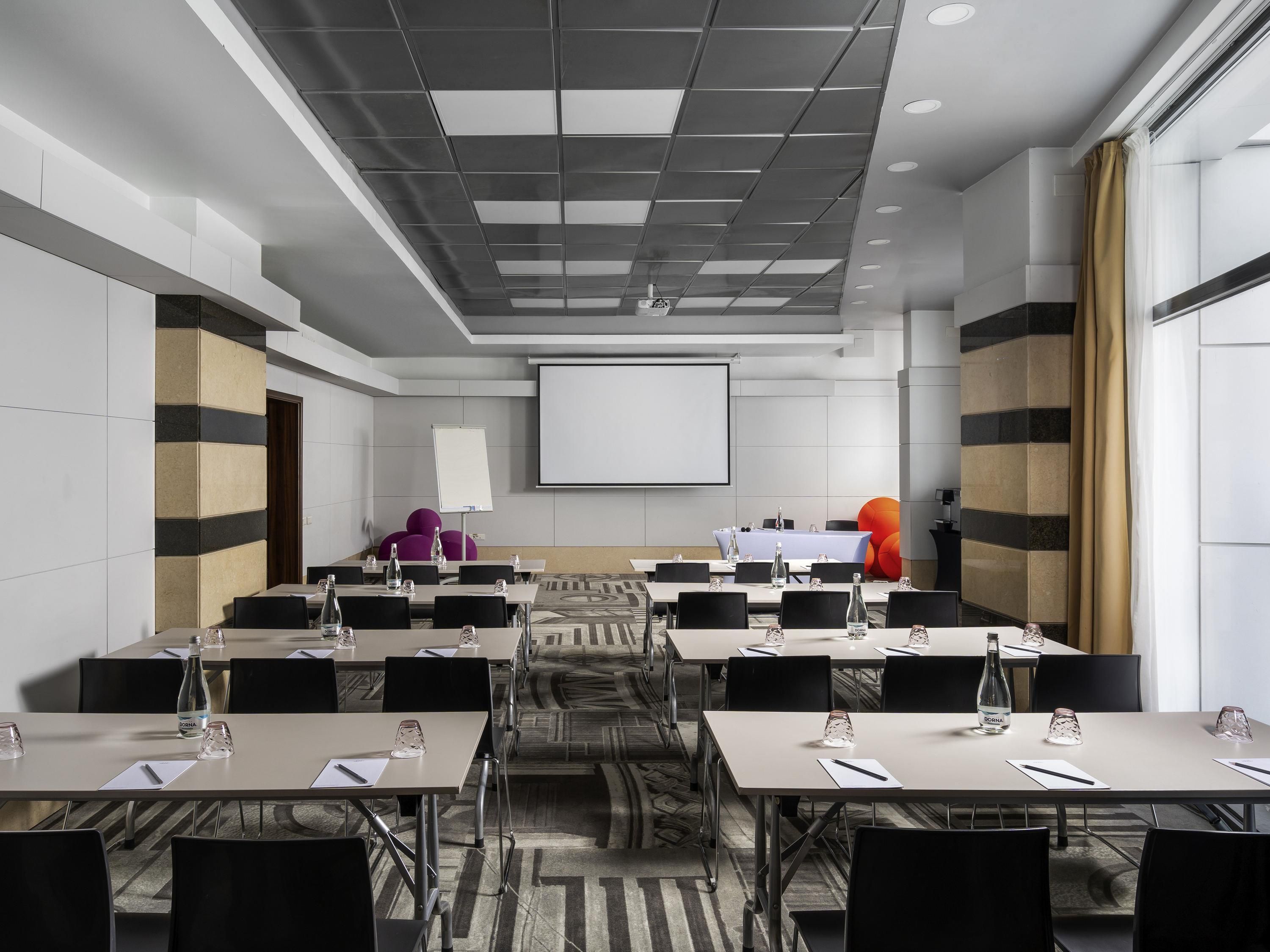 A conference room with tables, chairs, a projector screen, and a carpeted floor.