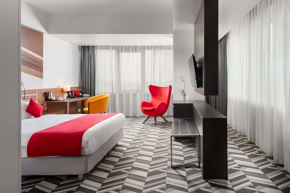 A hotel room with a bed, a desk, two chairs, a TV, and a large window with curtains.