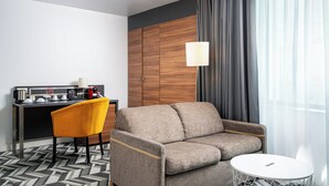 Una habitación de hotel moderna con sofá, una mesita redonda pequeña, una silla, un escritorio con cafetera y un armario.