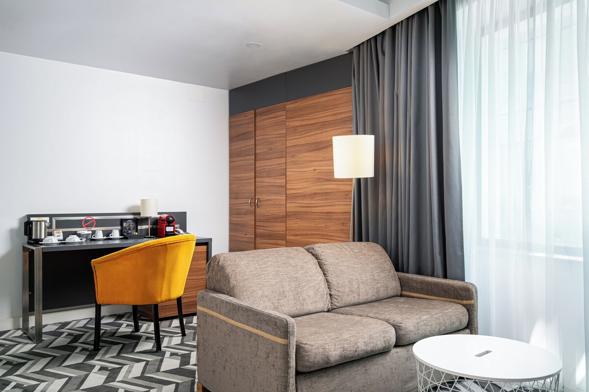 A modern hotel room with a sofa, a small round table, a chair, a desk with a coffee maker, and a wardrobe.