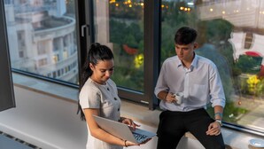 Dos personas en una oficina con una laptop y una taza de café, ventanas amplias con vista a la ciudad.