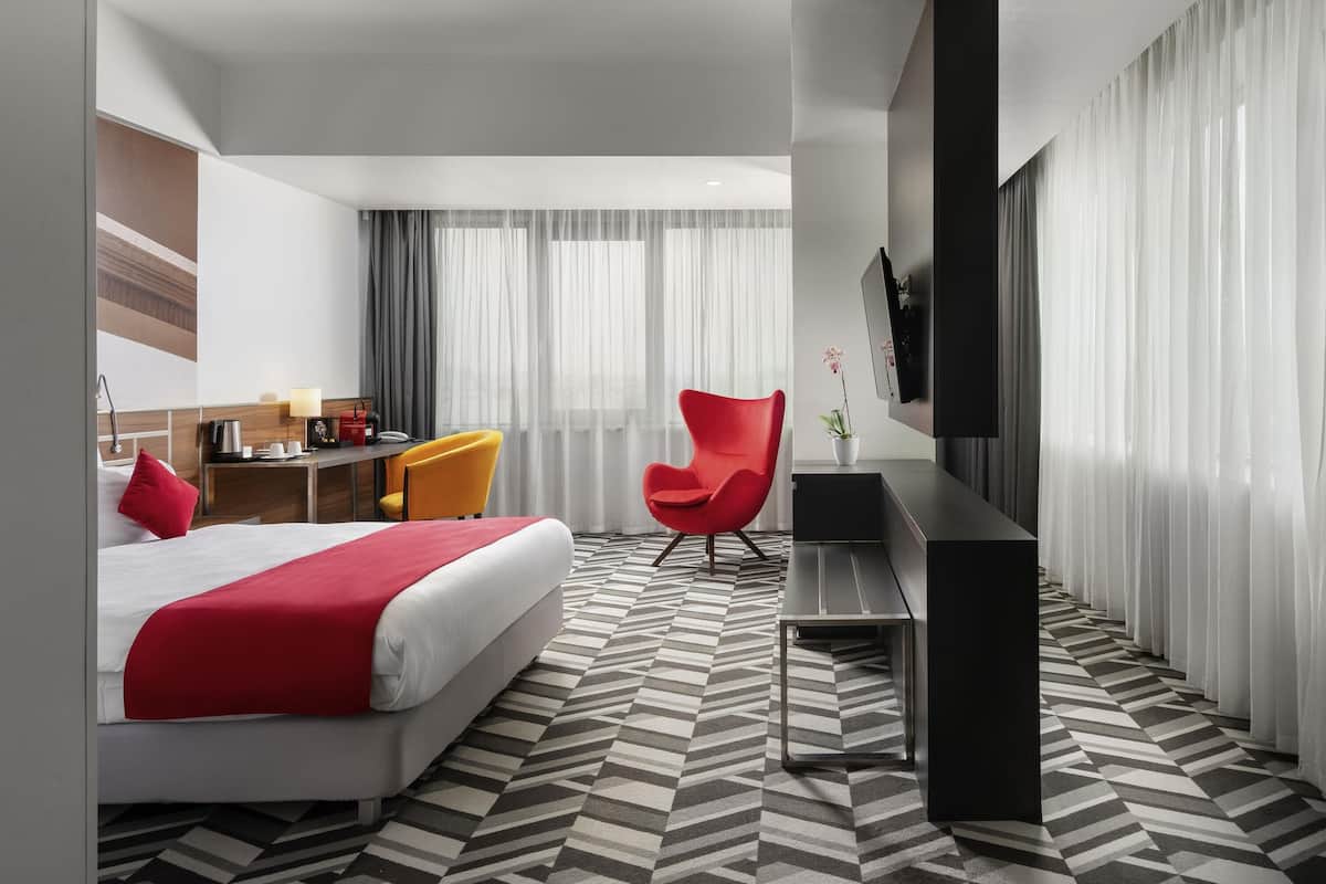Une chambre d’hôtel avec un lit, un bureau, deux chaises, une télévision et une plante en pot.