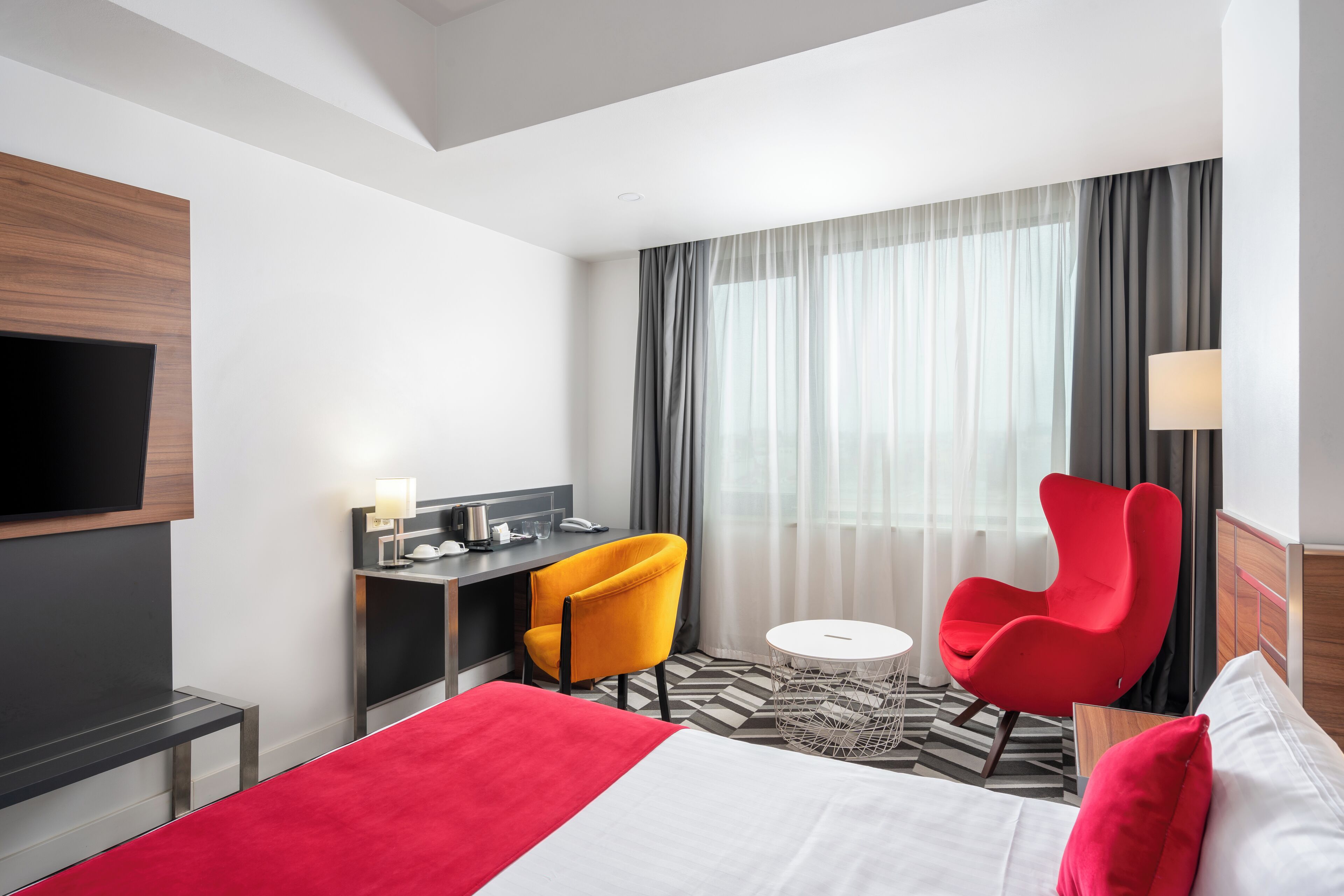 A hotel room with a bed, a desk, two chairs, a TV, and a window with curtains.