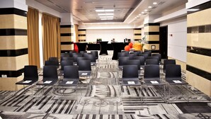 Un salón de conferencias con filas de sillas negras, una alfombra estampada y un mostrador de recepción al fondo.