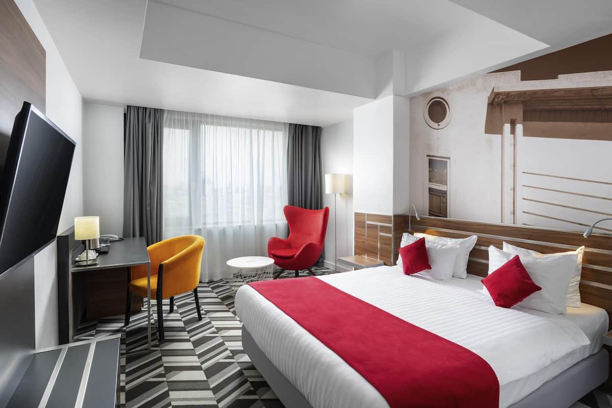 Une chambre d’hôtel avec un grand lit, une télévision, un bureau avec une chaise et une fenêtre avec des rideaux.