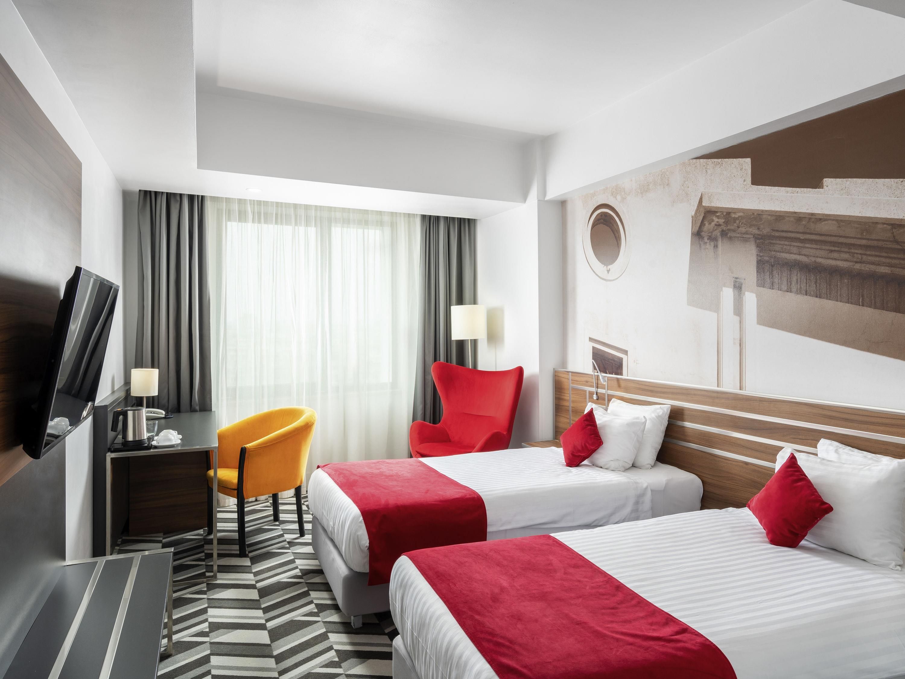 A hotel room with two beds, a TV, a desk, a chair, and a large window with curtains.