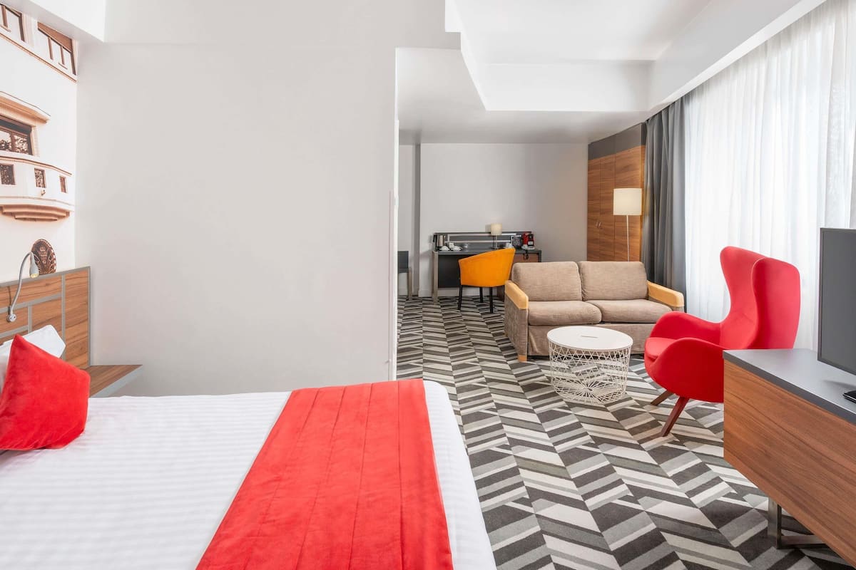 A hotel room with a bed, a sofa, a desk with a TV, and a dining area.