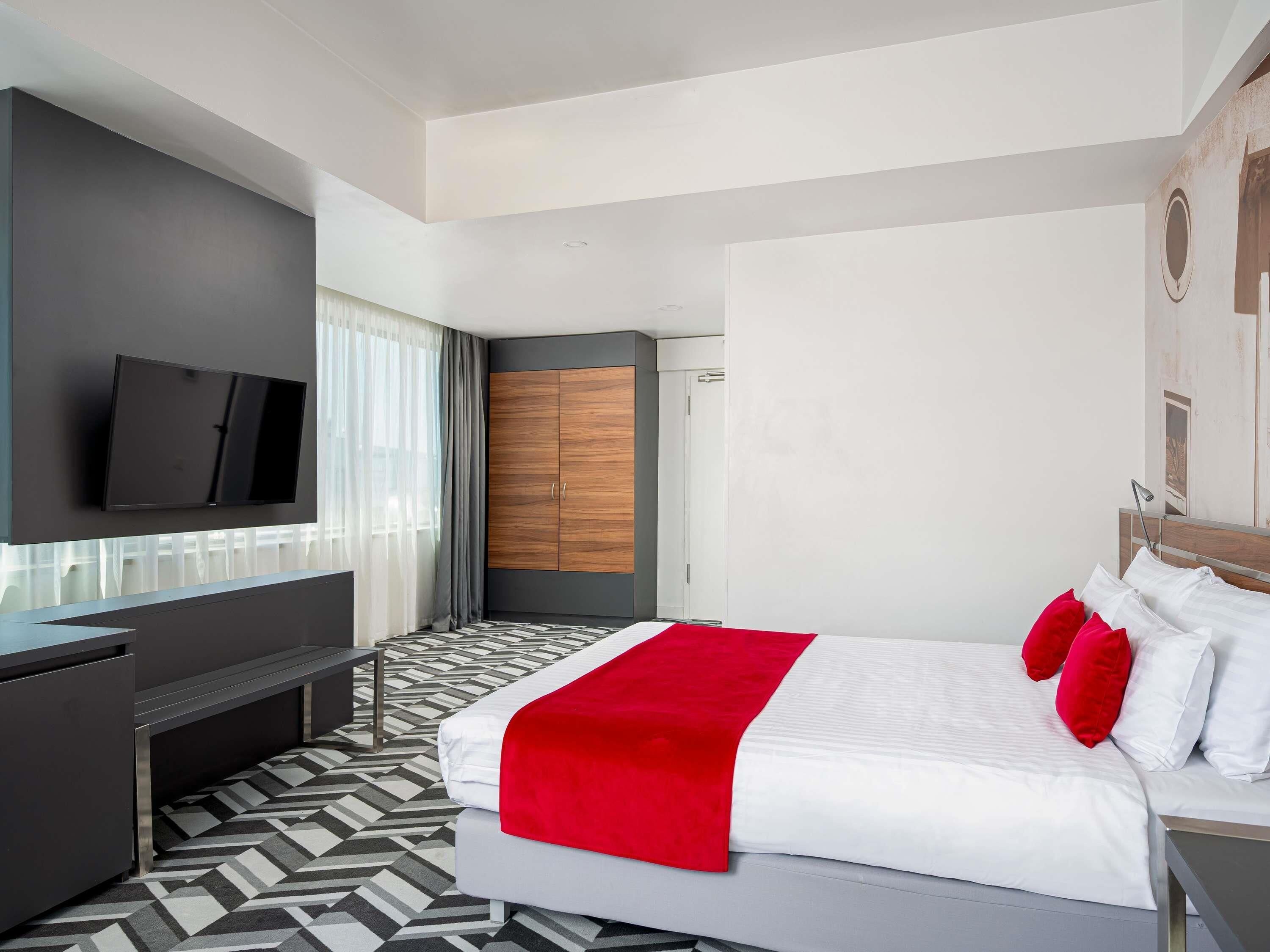 A modern hotel room with a bed, a TV, a wardrobe, and a desk.