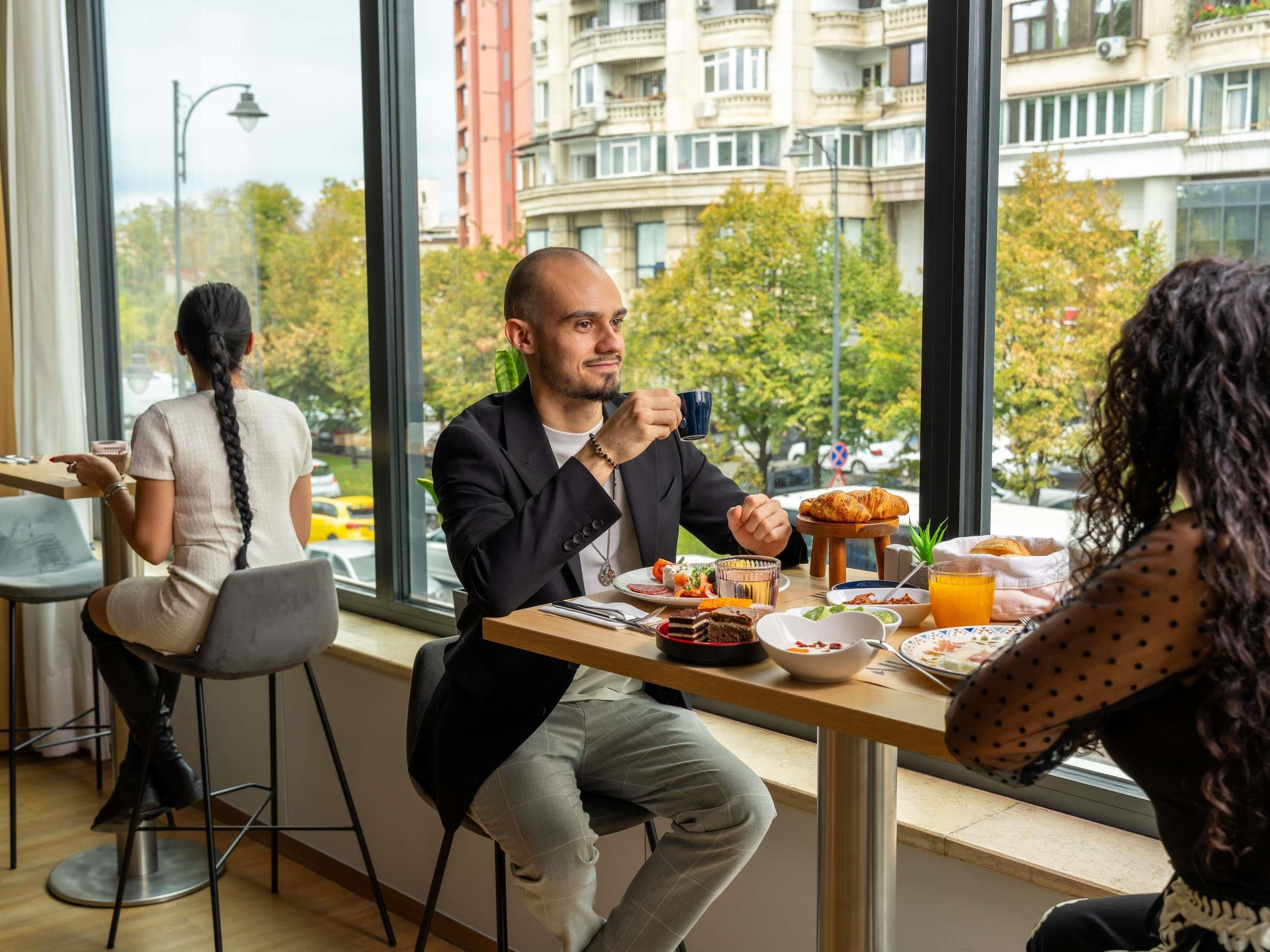 A dining area with a view of a cityscape, featuring a table with food and drinks, and a window overlooking the street.