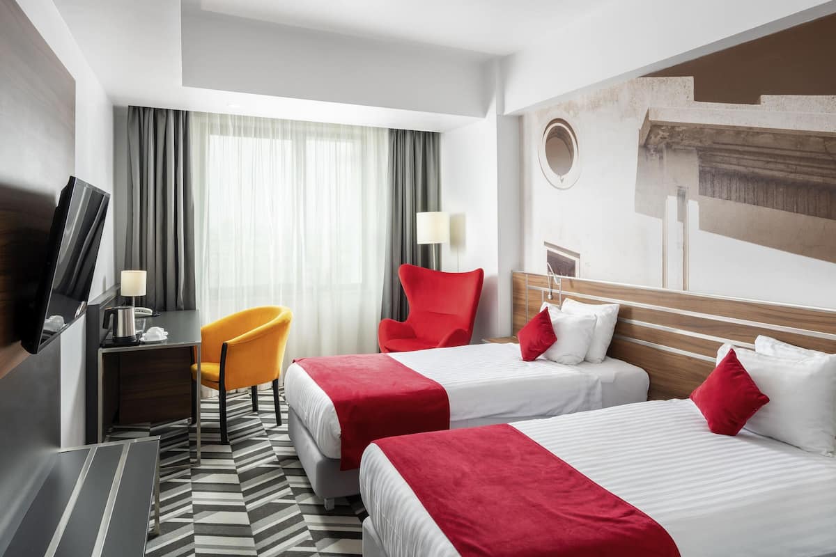 Une chambre d’hôtel avec deux lits, une télévision, un bureau, une chaise et une grande fenêtre avec des rideaux.