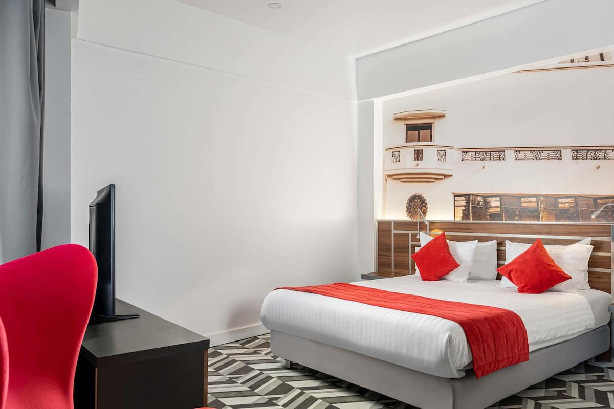 Une chambre d’hôtel moderne avec un grand lit, un fauteuil rouge, un téléviseur à écran plat et un sol à motifs.