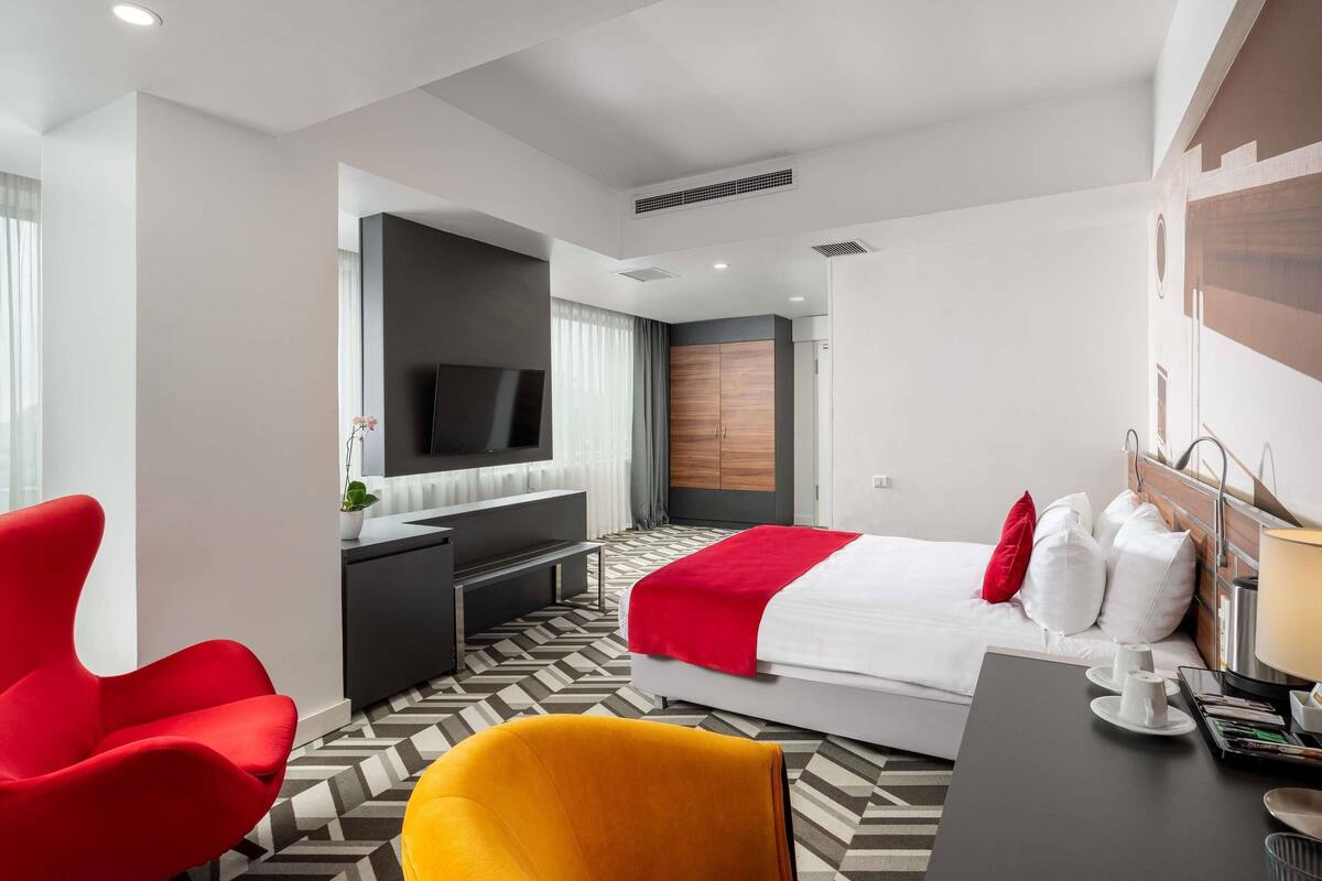 Une chambre d’hôtel moderne équipée d’un lit, d’un bureau, d’une télévision et d’un fauteuil rouge.