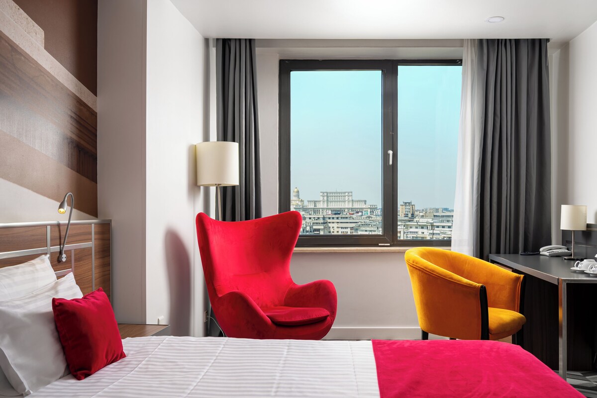 A hotel room with a bed, a red armchair, a yellow armchair, a desk with a lamp, and a window with a city view.