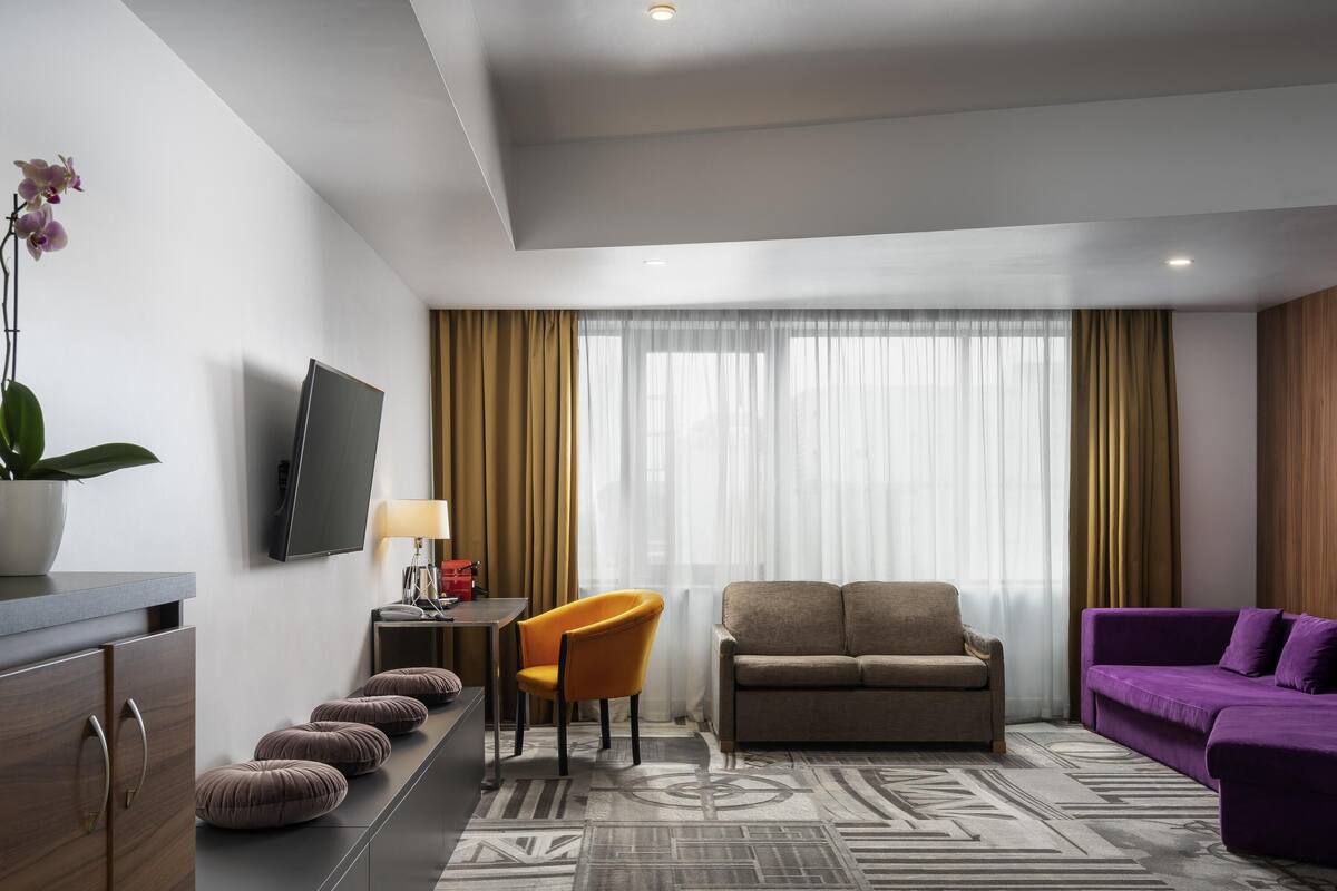 Una habitación de hotel moderna con sofá, un sofá morado, un escritorio con lámpara, televisión y un ventanal con cortinas.