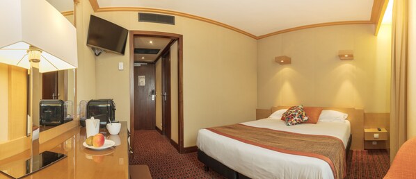Habitación de hotel con cama, mesita de noche, lámpara, televisor y un pequeño escritorio con cafetera y fruta.