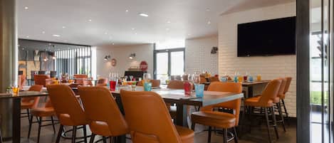 A modern dining area with multiple tables and orange chairs, a flat-screen TV mounted on the wall, and a brick wall design.