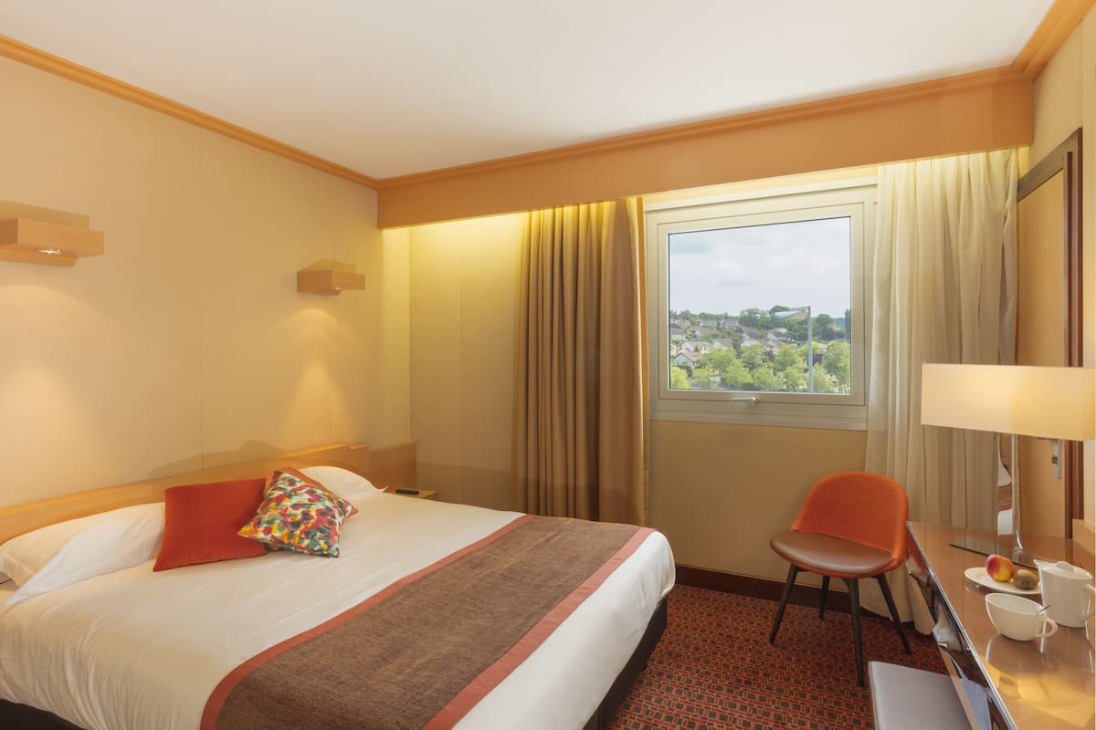 Ein Hotelzimmer mit Bett, Stuhl, Schreibtisch mit Lampe, Tasse und Blick ins Freie.