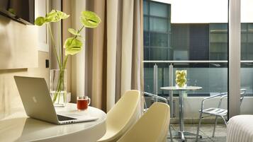 A modern hotel room with a laptop on a round table, a glass of beverage, and a vase with green leaves. There is a balcony with a glass railing and a small table with a vase of yellow flowers.