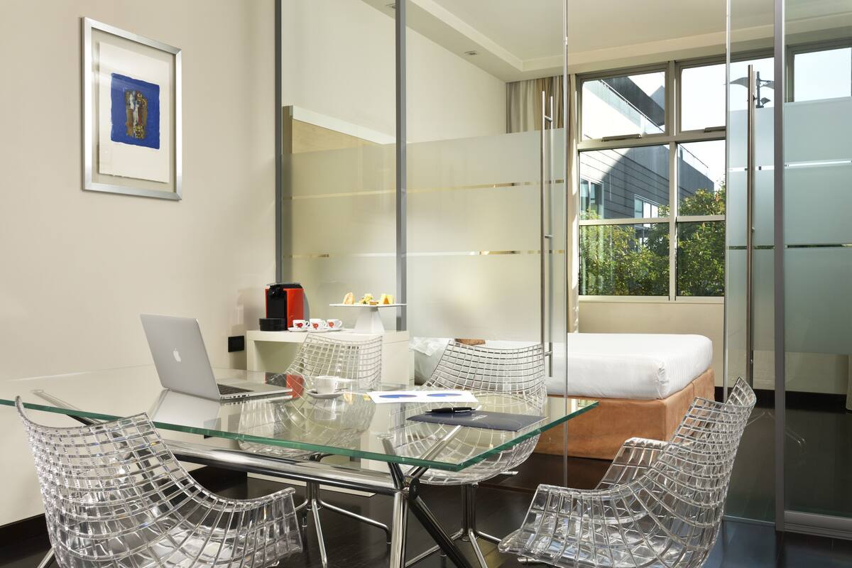 Habitación de hotel moderna con mesa de vidrio, sillas de estructura metálica transparente, una computadora portátil, una cama y un ventanal con vistas al exterior.