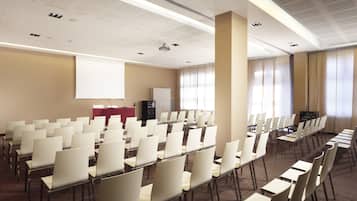A conference room with rows of chairs, a podium, a projector screen, and a large window with curtains.