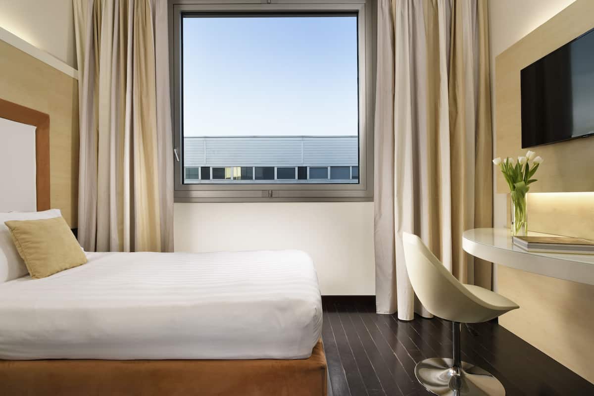 Una habitación de hotel con cama, escritorio, silla, televisión y ventana con cortinas.