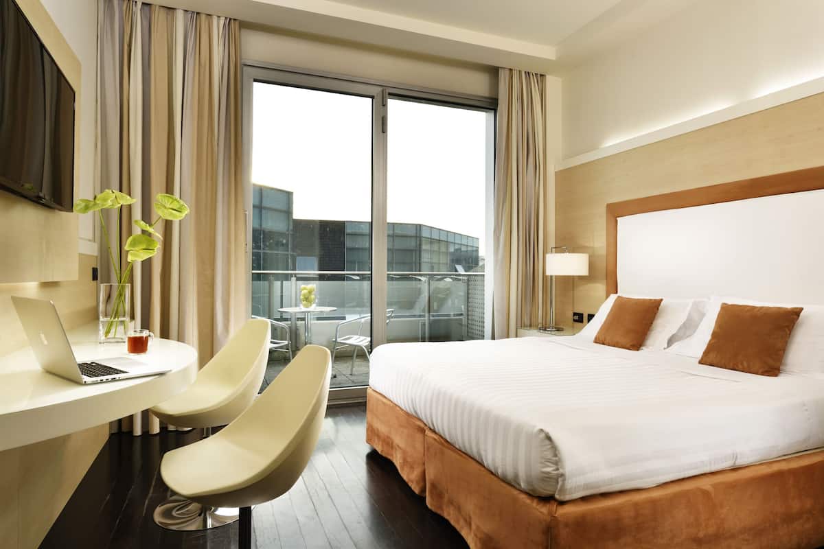 Habitación de hotel con cama, escritorio y silla, una computadora portátil, un florero con flores y vistas a un edificio a través de la ventana.