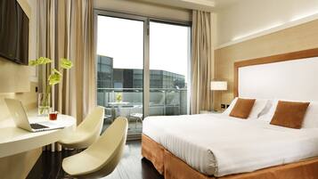 Habitación de hotel con cama, escritorio y silla, una computadora portátil, un florero con flores y vistas a un edificio a través de la ventana.