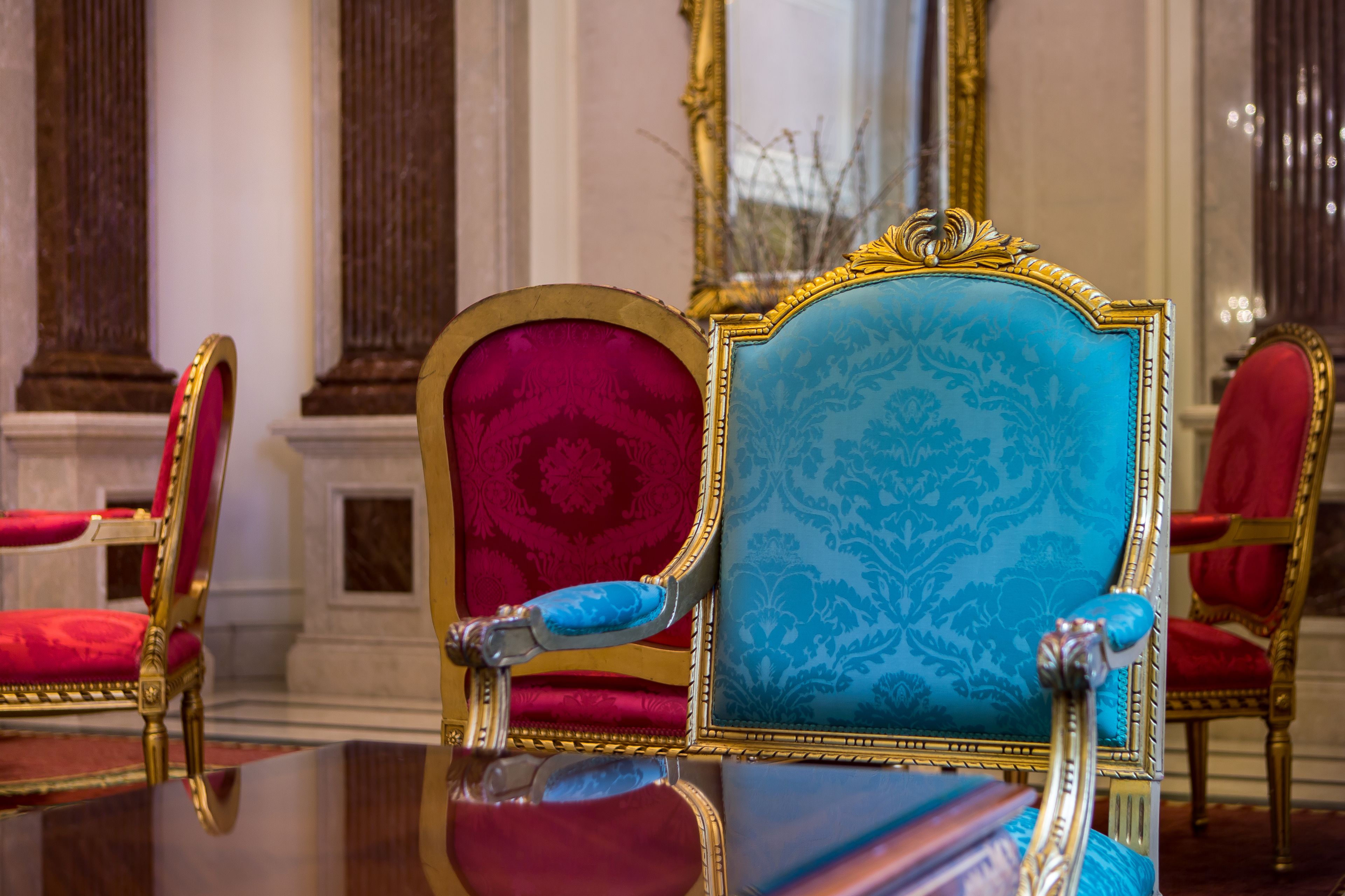 Una habitación lujosa con muebles ornamentados, incluyendo una butaca azul y roja con detalles dorados, y un espejo grande que refleja la decoración de la habitación.