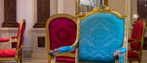 Una habitación lujosa con muebles ornamentados, incluyendo una butaca azul y roja con detalles dorados, y un espejo grande que refleja la decoración de la habitación.