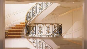 A grand spiral staircase with ornate black and gold railings, leading to a landing with a polished floor and a reflection of the ceiling.