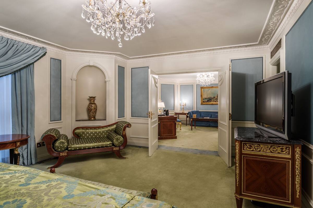 A spacious hotel room with a large bed, a sitting area with a sofa, a television, and a chandelier.