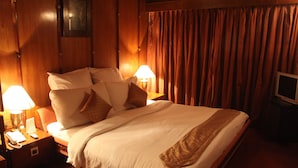 A neatly made bed with white linens and brown accents, two bedside lamps, a nightstand with a telephone and remote control, and a television on a stand to the right. - Saj Luciya (Thiruvananthapuram)