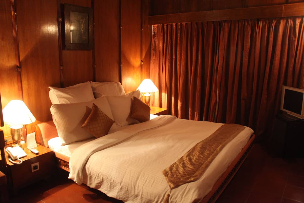 Una cama cuidadosamente tendida con ropa de cama blanca y detalles marrones, dos lámparas de noche, una mesita de noche con un teléfono y control remoto, y un televisor sobre un mueble a la derecha.