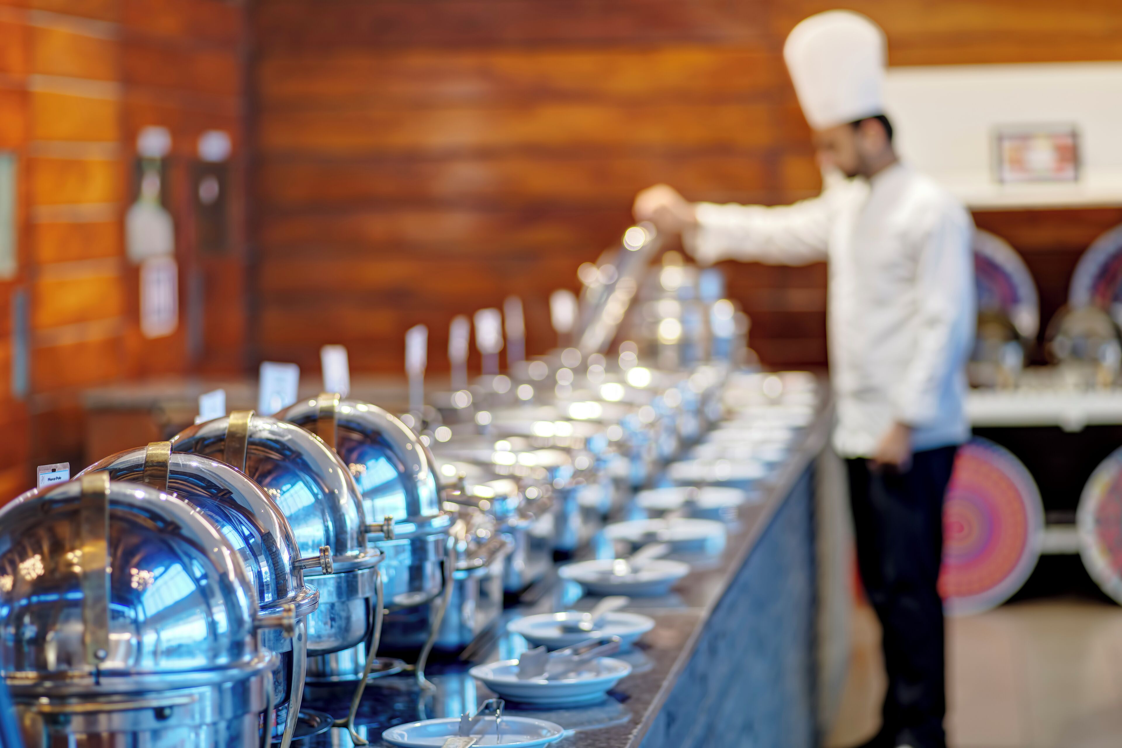 A buffet line with covered food containers and a person in chef attire serving food.