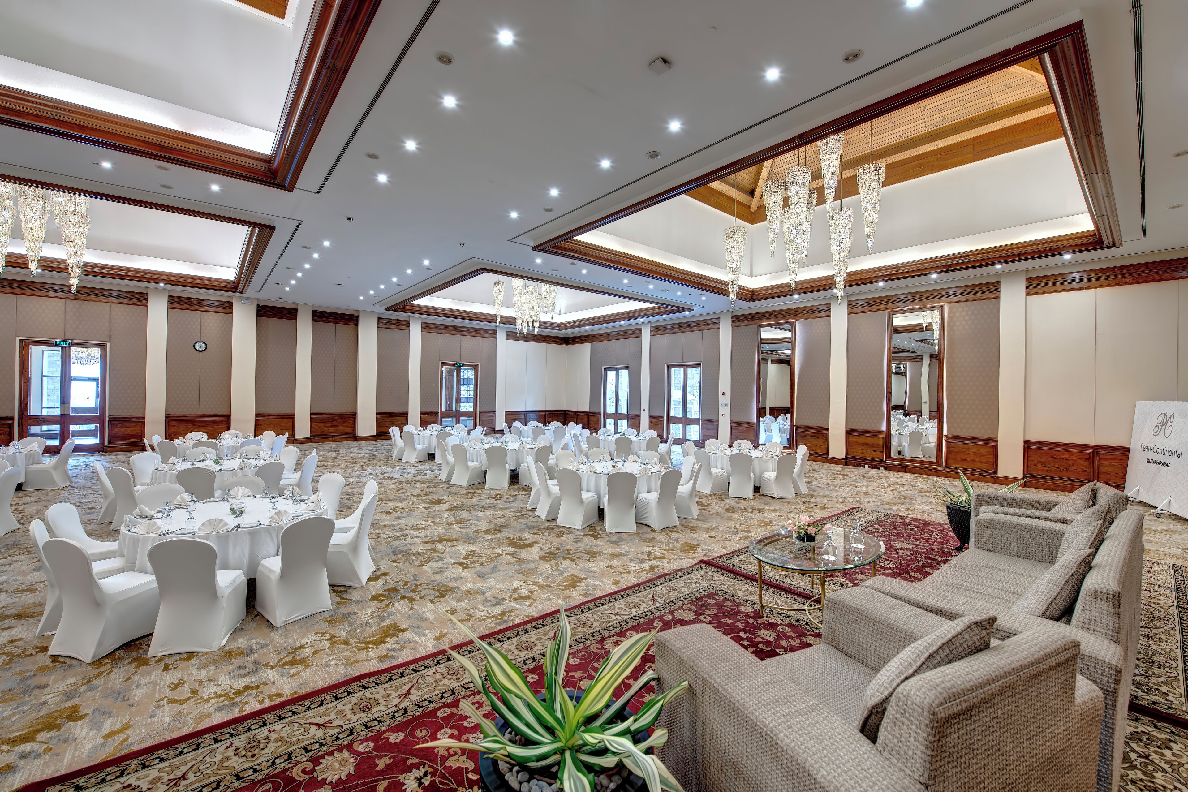 A spacious banquet hall with round tables and white chairs, a large potted plant, and a sofa area.