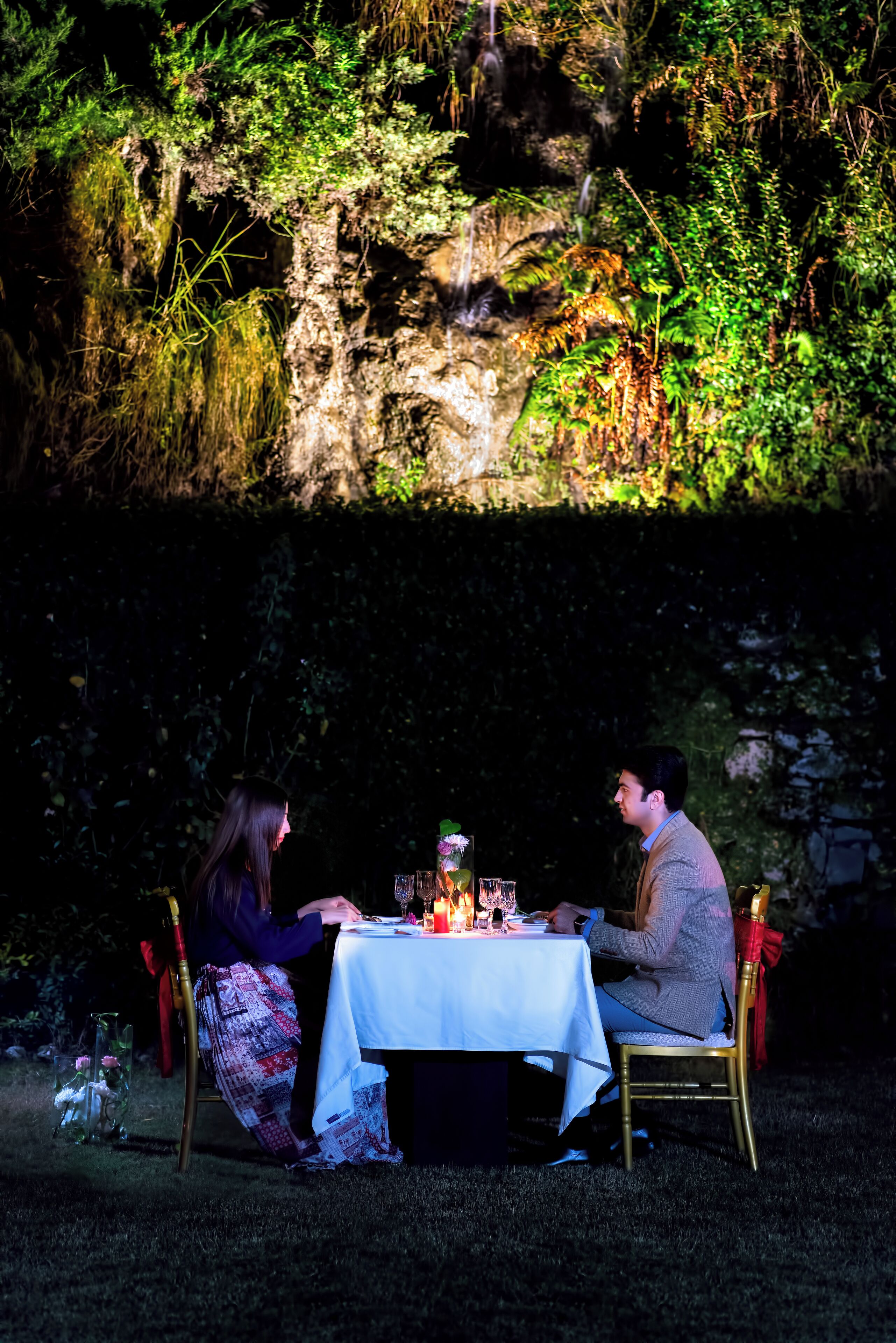 An outdoor dining table with a white tablecloth, surrounded by chairs, set against a backdrop of greenery and a rocky wall.