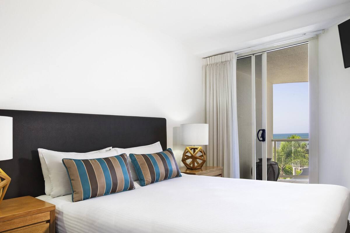 Una habitación de hotel con cama, mesitas de noche, lámpara y vista al mar.