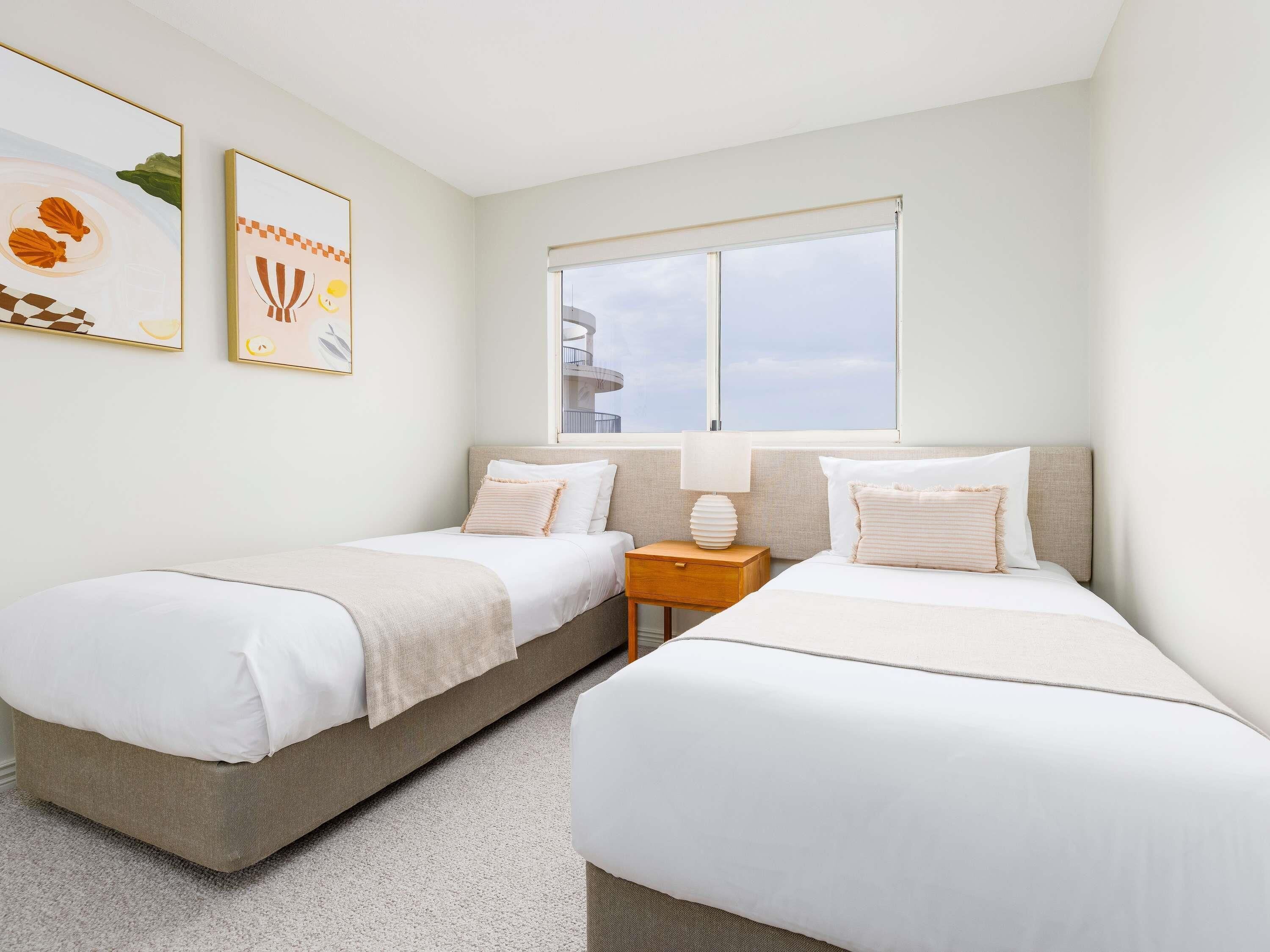 A hotel room with two single beds, a window with a view, and two framed artworks on the wall.