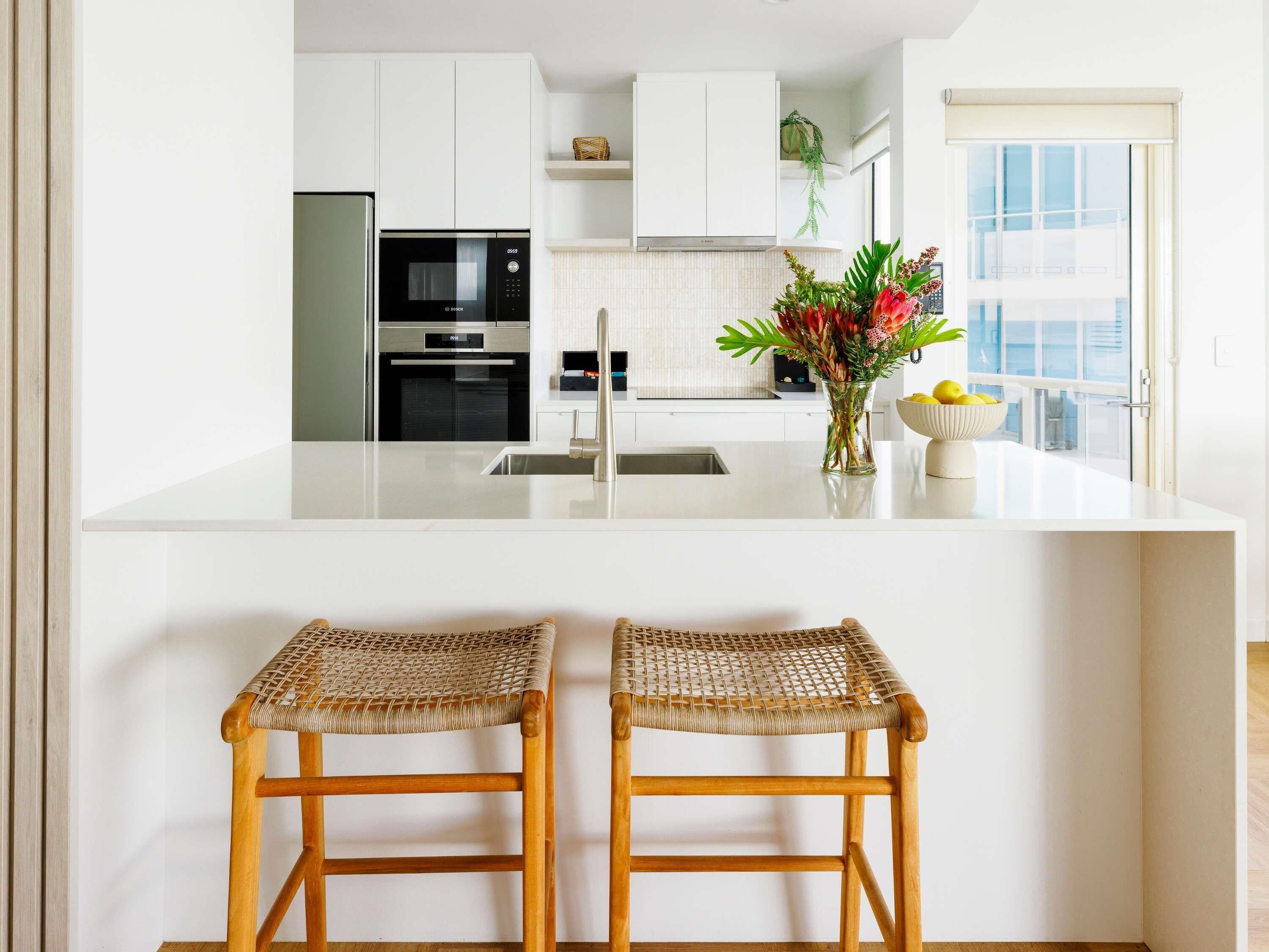 A modern kitchen with white cabinetry, a built-in microwave, and a large window. There are two wicker bar stools and a vase with flowers on the counter.