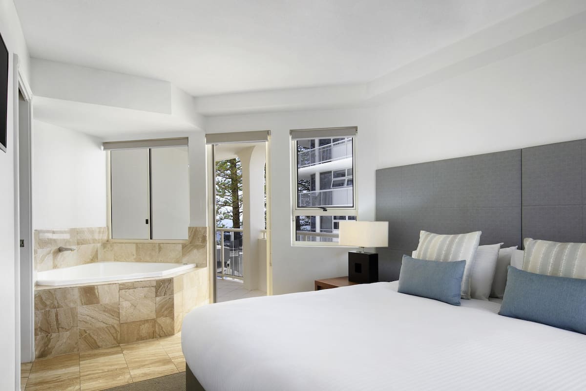 Una habitación de hotel moderna con una cama grande, baño independiente con bañera, balcón con vistas al edificio y mesita de noche con lámpara.