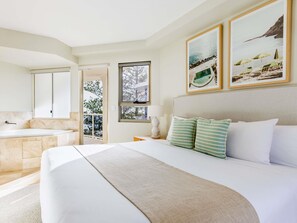 Habitación de hotel con una cama grande, baño con bañera y balcón con vistas a árboles.