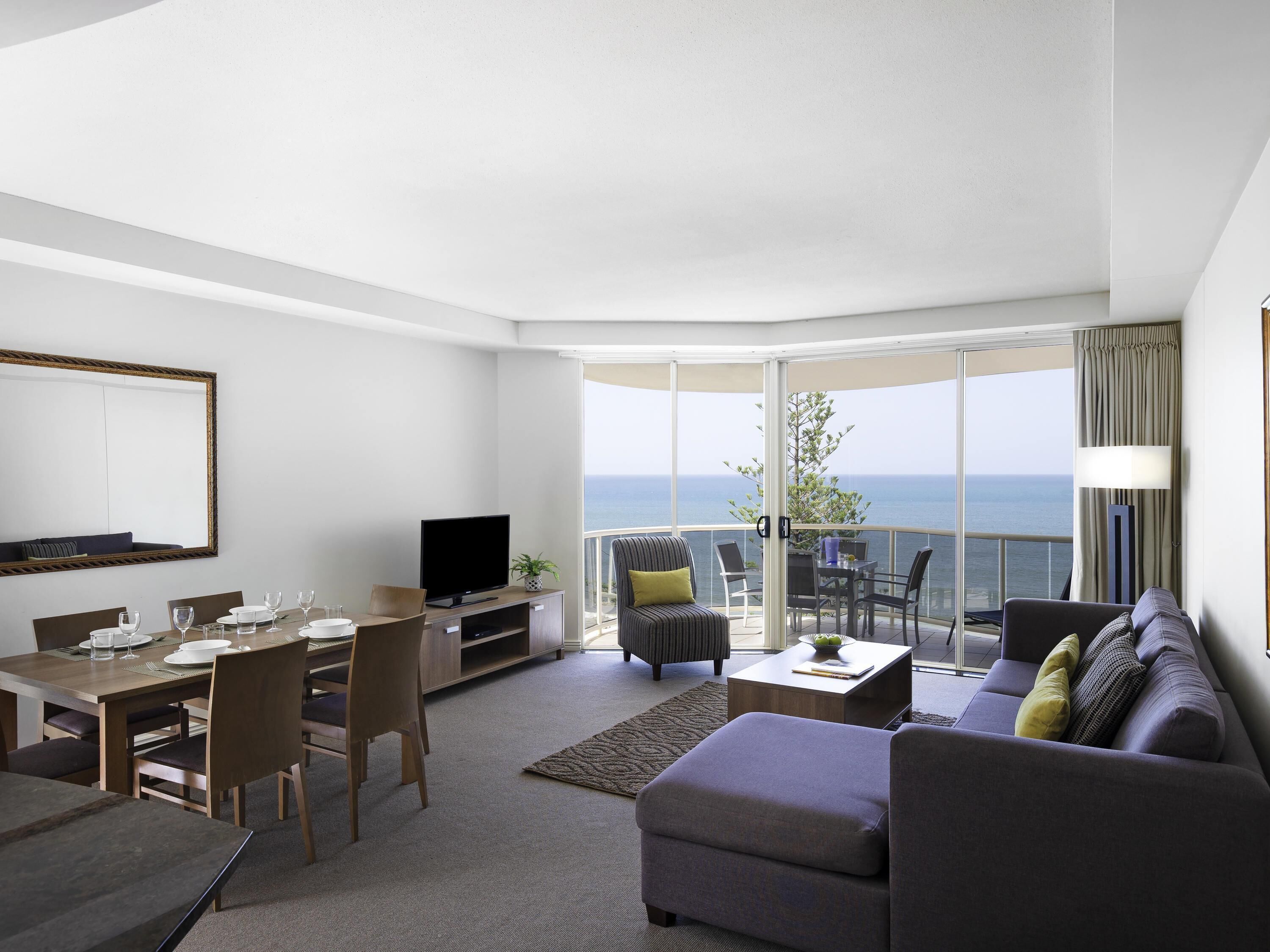 Un salon moderne comprenant un coin repas, un téléviseur à écran plat et un balcon offrant une vue sur l’océan.