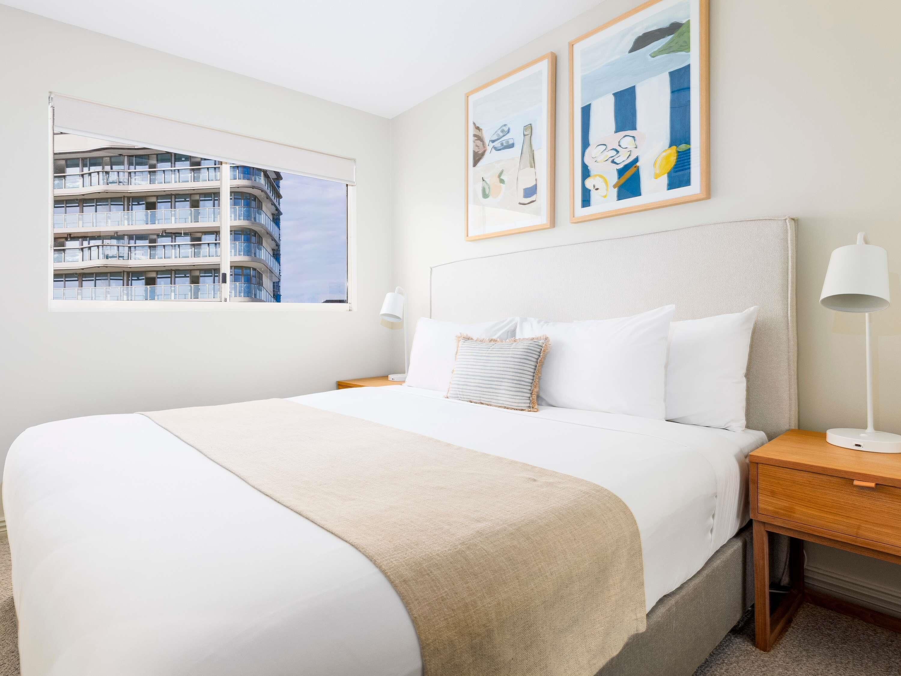 A hotel room with a large bed, a bedside table with a lamp, a window with a view of a building, and two framed pictures on the wall.