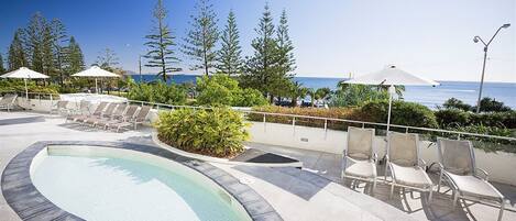 A pool area with lounge chairs and umbrellas, surrounded by trees and overlooking the ocean.