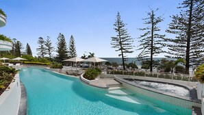 A swimming pool with a curved design, surrounded by lounge chairs and umbrellas, with tall trees and a clear sky in the background. - Mantra Sirocco Resort (Mooloolaba)