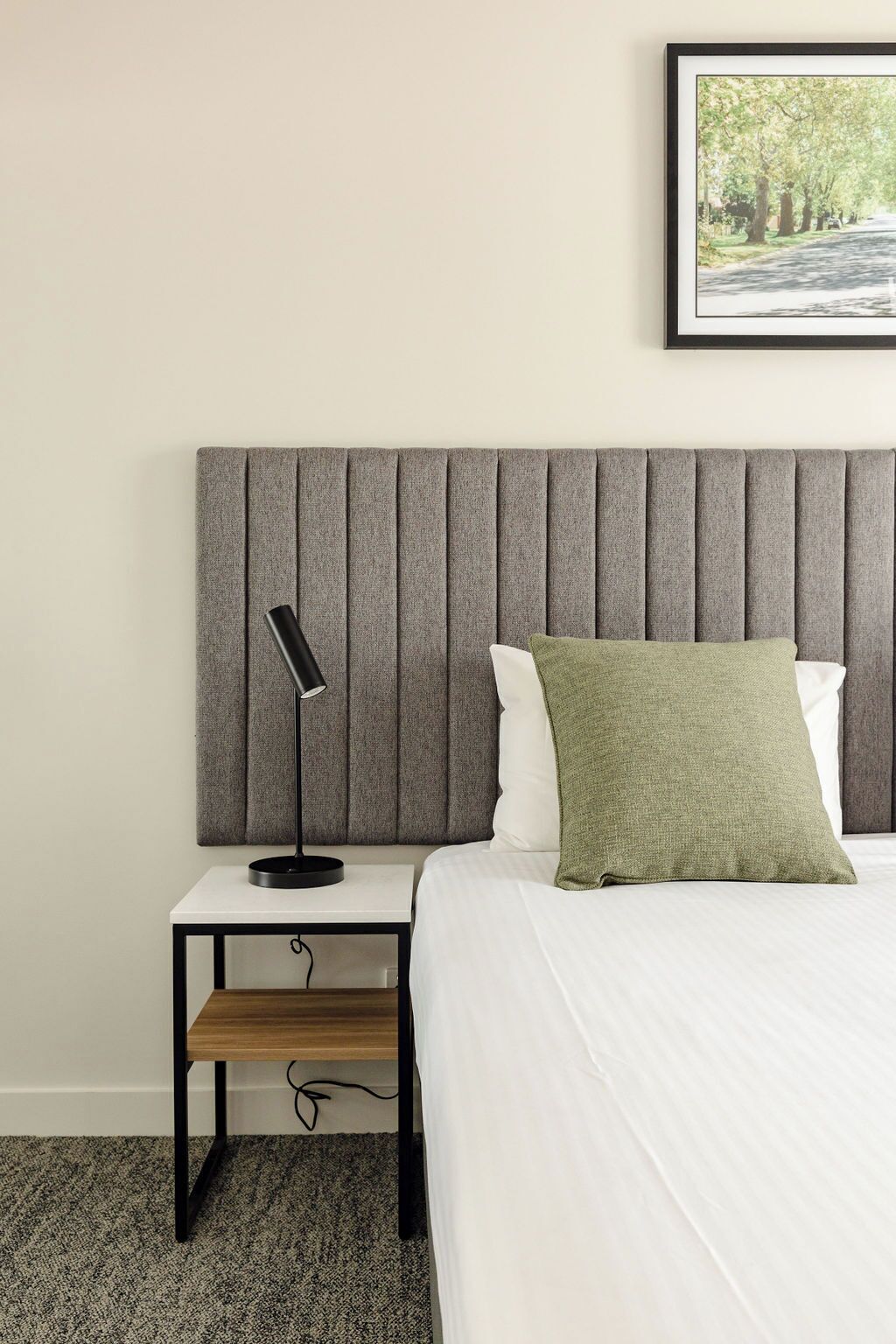 A neatly arranged hotel room with a bed, a bedside table, a lamp, and a framed picture on the wall.